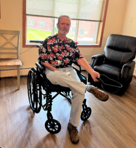 A man using a wheelchair in an apartment, with a chair and window behind him. 