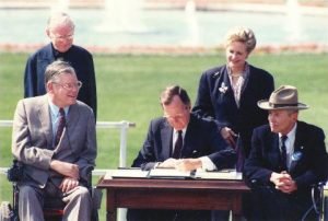 Group of people in suits surrounding a desk while president signs a paper. 